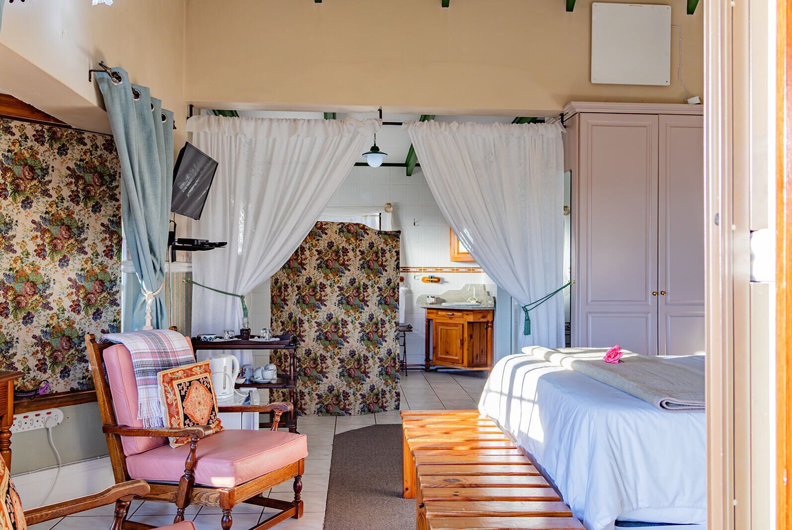 A cozy bedroom with a wooden bed, a desk with a chair, a small kitchenette, and a bathroom visible through a curtain.