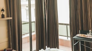 Une chambre d’hôtel avec un balcon, de grandes fenêtres avec des rideaux, une table à manger avec une assiette et des verres, et une petite étagère avec une figurine.