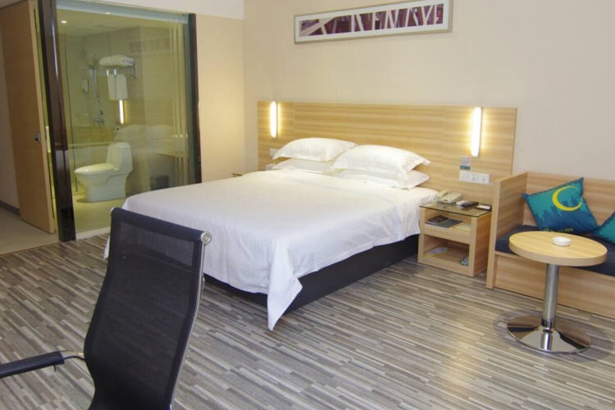 A hotel room with a bed, a chair, a small table, a nightstand, and a glass-enclosed shower area.