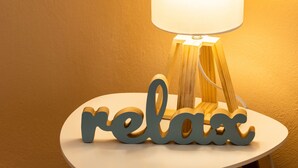 A table lamp with a white shade and a wooden base, placed on a small white table with the word "relax" spelled out in blue letters. - Zennova 5 Antigonidon Sq (Thessaloniki)