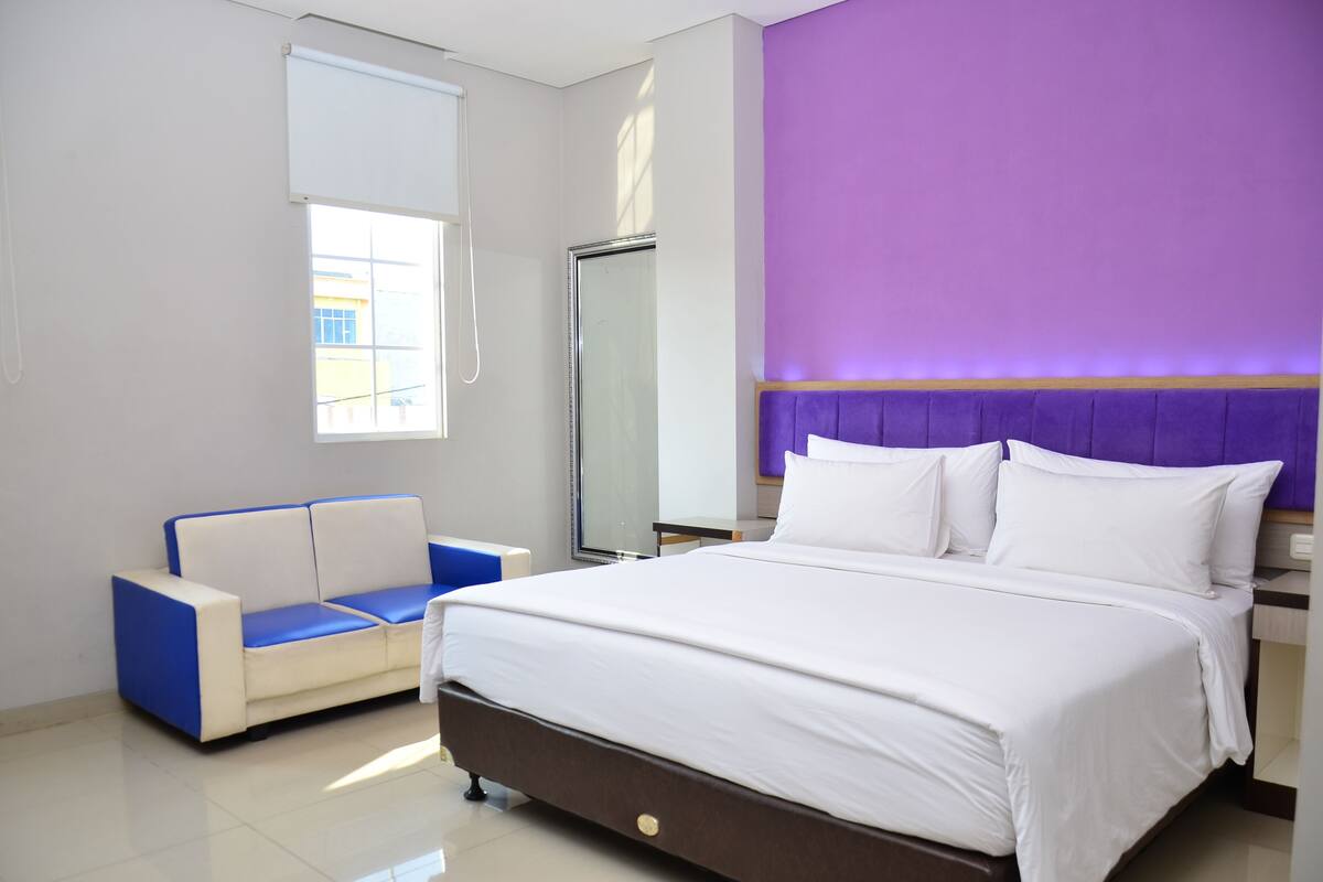 A modern hotel room with a large bed, a blue and white sofa, a large mirror, and a purple accent wall.
