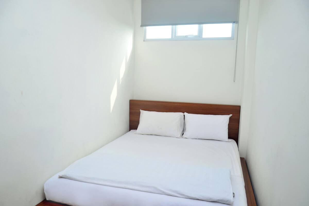A small, single-bed room with a wooden headboard, white bedding, and a window with a blind.