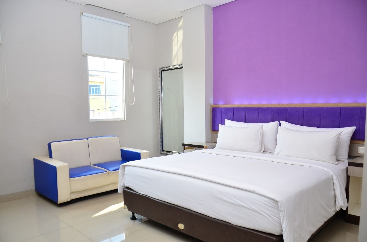 A hotel room with a bed, a blue and white sofa, a window, and a purple accent wall.