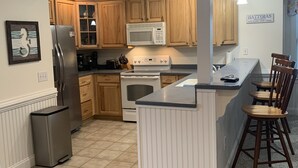 A kitchen with wooden cabinets, a stainless steel refrigerator, a microwave, a stove, and a small trash can. - SeaToo Waterfront Condo (Hatteras)