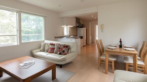 A modern living room with a sofa, coffee table, and dining table set for two. A kitchen is visible in the background. - Sakura (Kutchan)