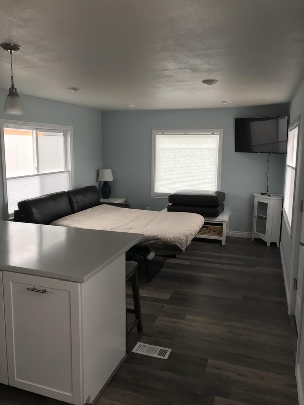 A compact, modern room with a kitchenette, a bed, a desk, and a TV mounted on the wall.