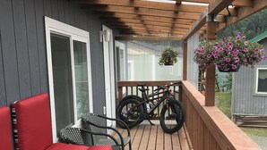 Een overdekt terras met een houten dek, rode, beklede zitplaatsen, een fiets en een bloemenmand.