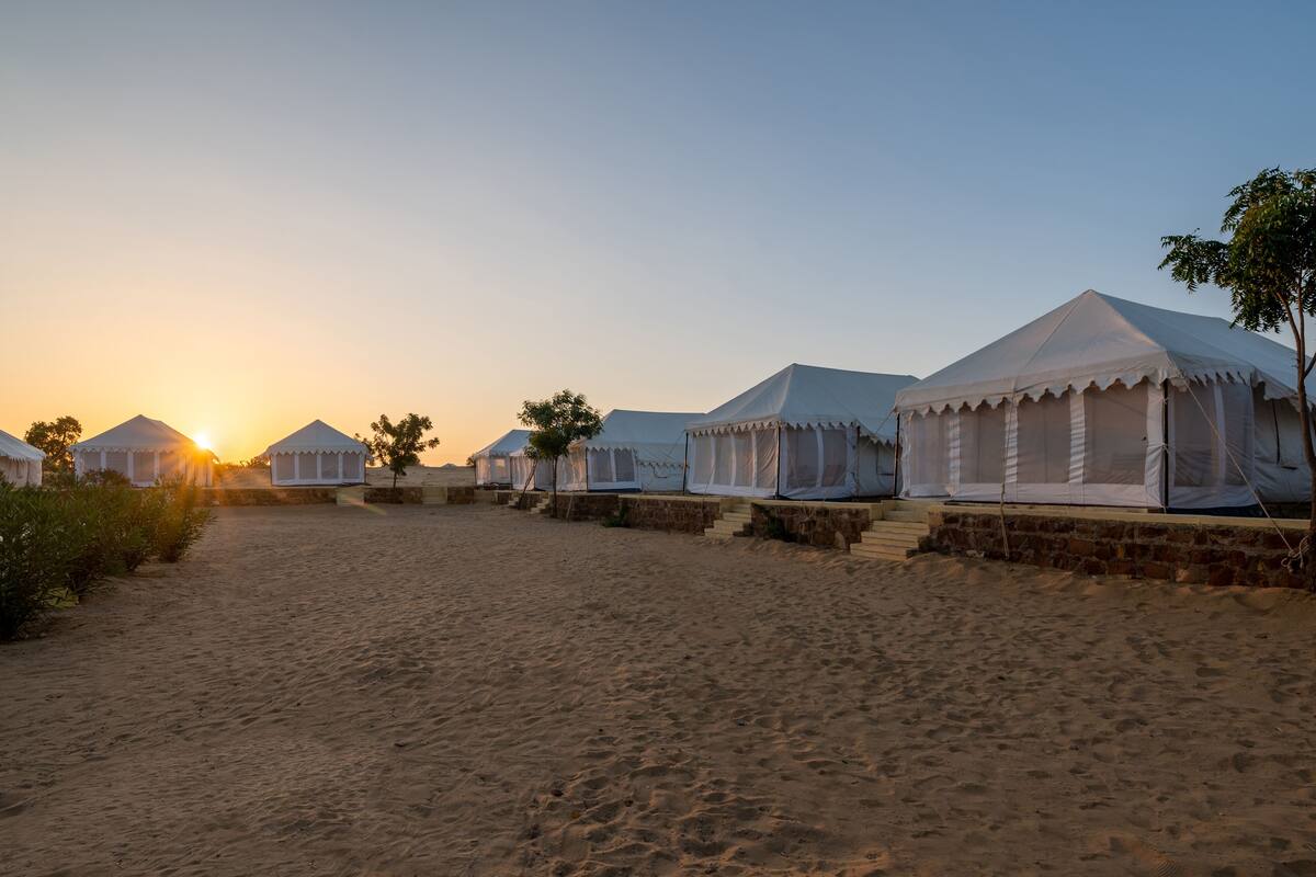 Eine Reihe weißer Zelte mit Backsteinunterbau, auf einem Sandstrand bei Sonnenuntergang.