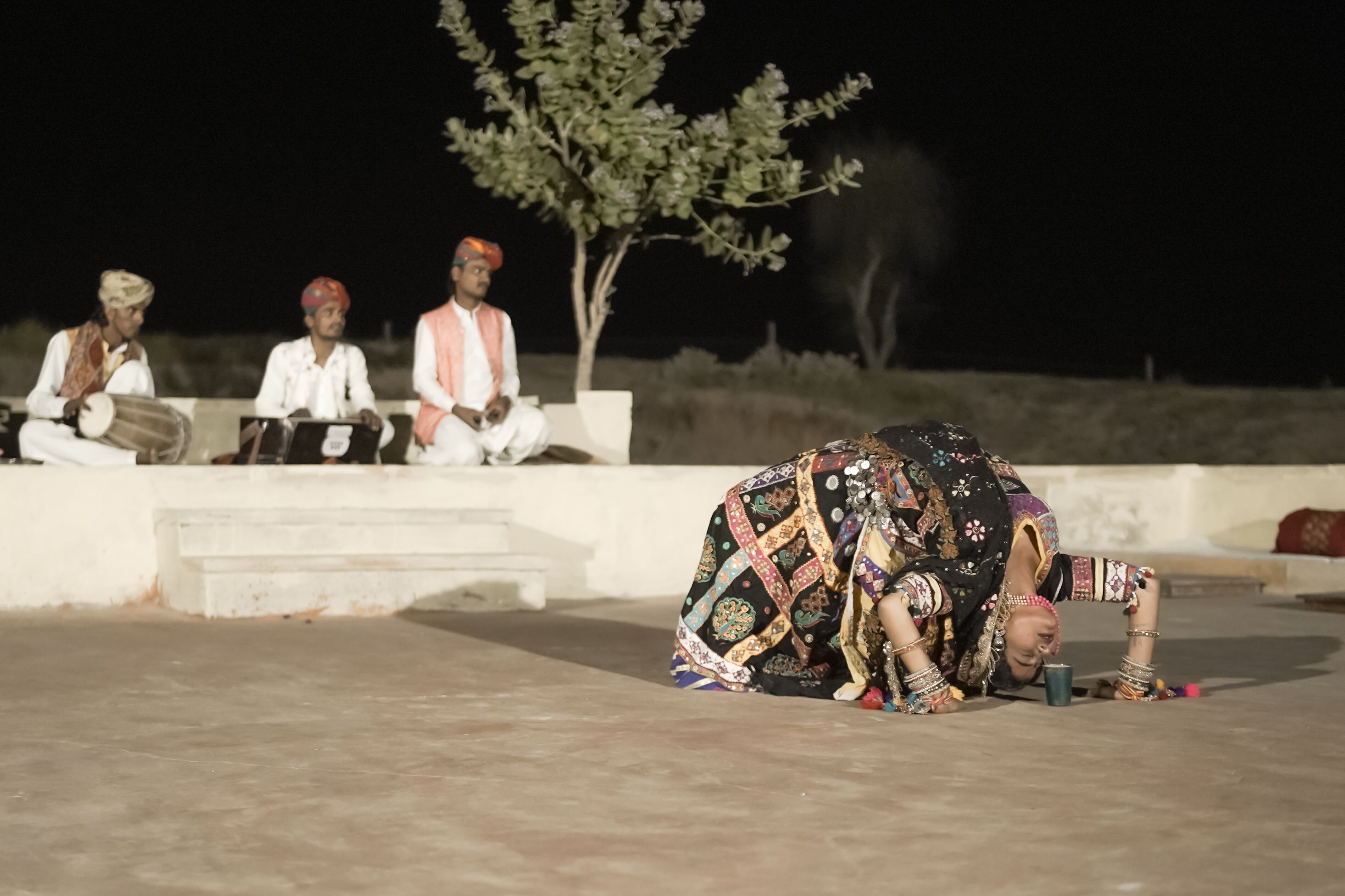 A person performing a traditional dance on a stage with a tree and a group of seated individuals in the background.