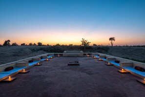 A rooftop terrace with seating areas and candles, overlooking a desert landscape at sunset.
