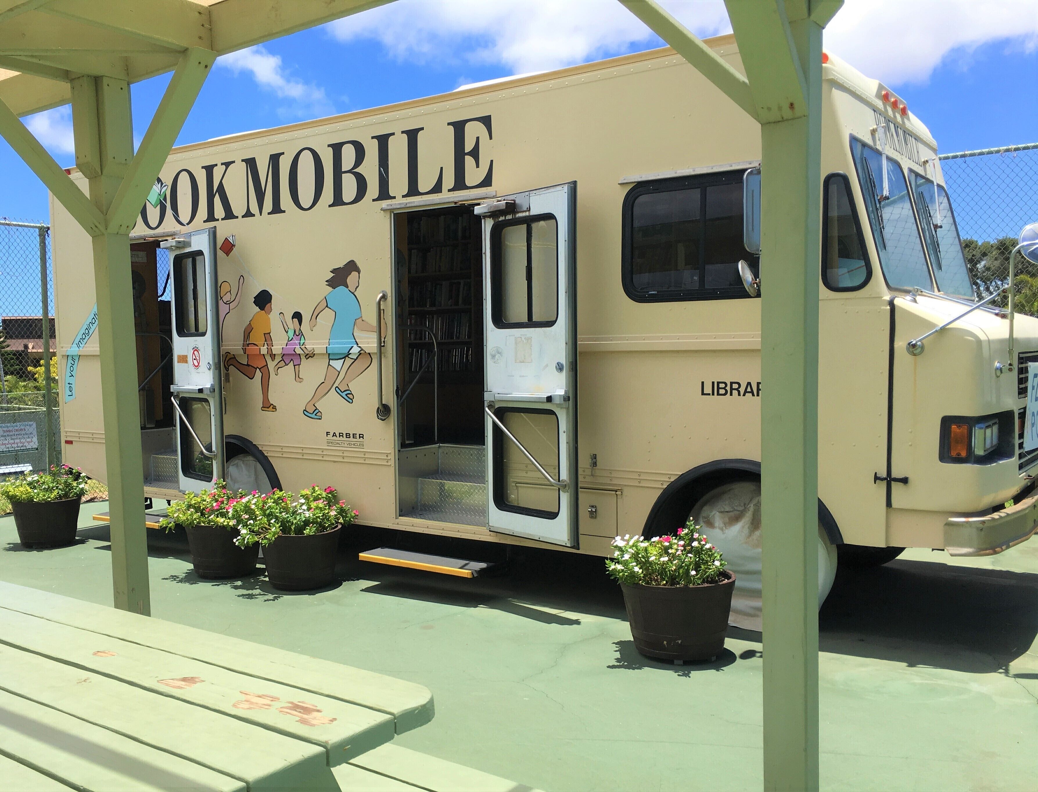 A mobile library with a 'Farber' logo, parked under a shelter with potted plants and a picnic table.
