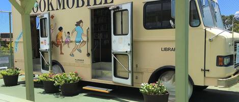 A mobile library with a 'Farber' logo, parked under a shelter with potted plants and a picnic table.