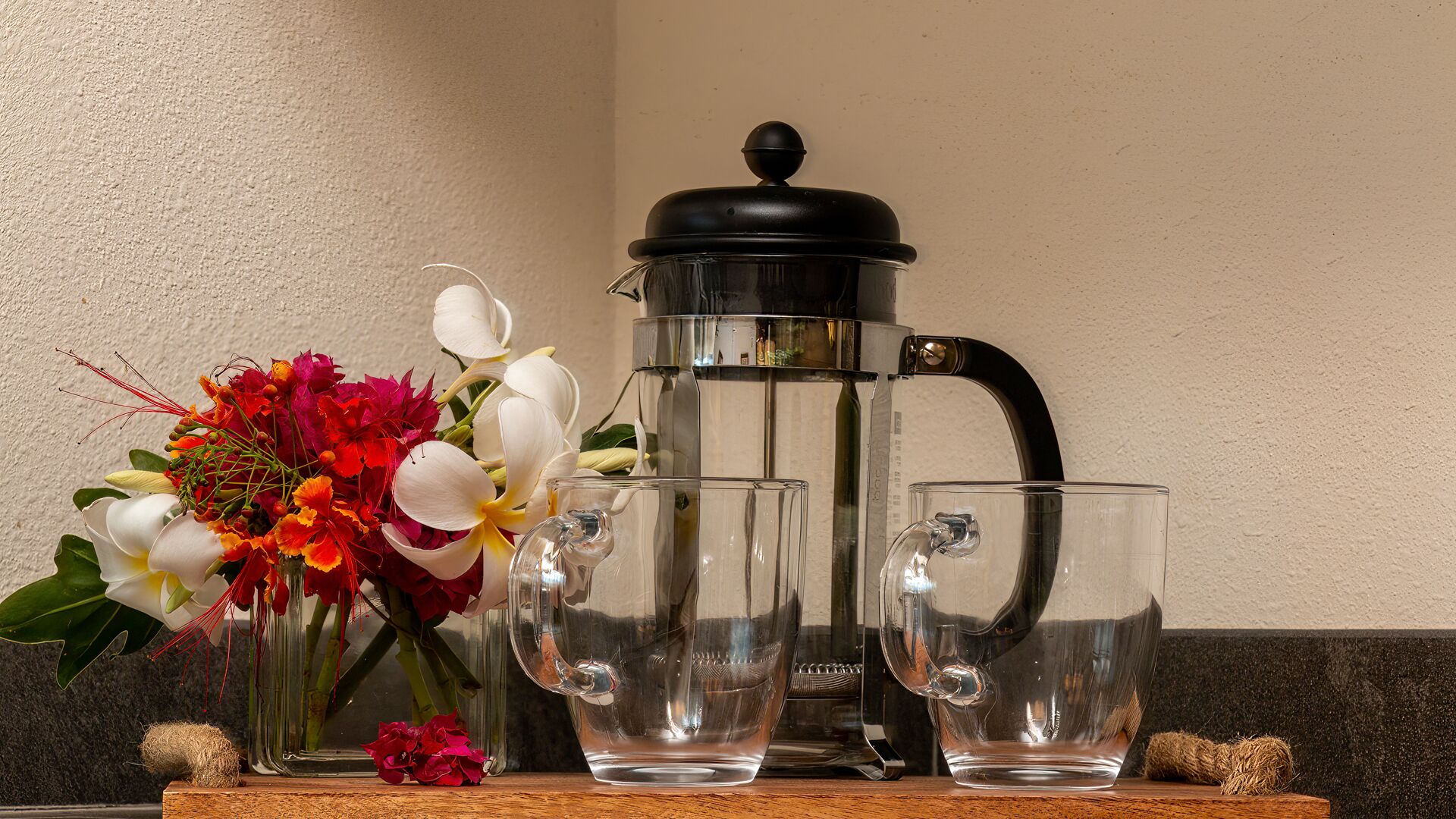 A coffee maker, two glasses, and a floral arrangement on a wooden surface.