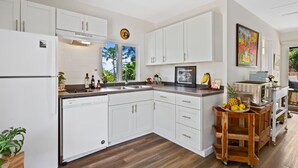 A modern kitchen with white cabinets, a dishwasher, and a microwave. There is a window with a view of greenery outside. - Reserve Your 5 STAR Big Island Get-a-way ⭐⭐⭐⭐⭐ (Waikoloa Village)