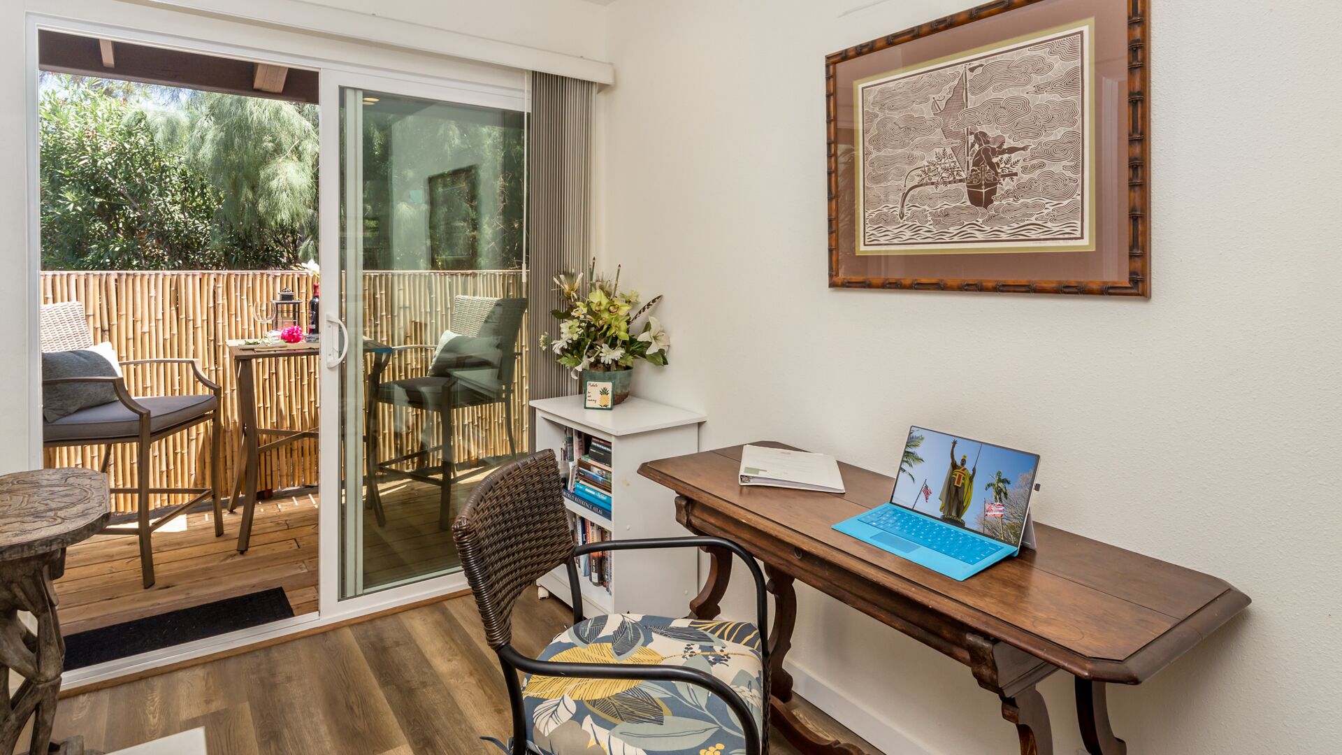 A home office with a laptop on a desk, a chair, a bookshelf, and a sliding glass door leading to an outdoor patio with chairs and a table.