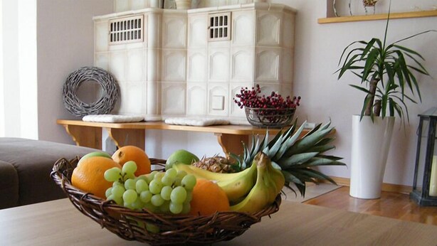 Una habitación con una mesa de madera, una cesta de mimbre con fruta, un armario blanco, una planta en maceta y un farol.