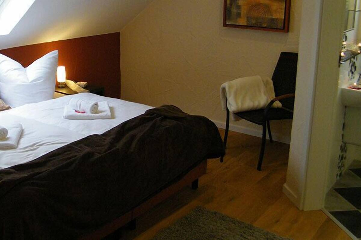 Ein Hotelzimmer mit Bett, zwei Kissen, einem Nachttisch, einem Sessel und einem kleinen Tisch mit Lampe.