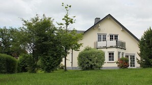 Una casa de dos pisos con balcón, rodeada de abundante vegetación y árboles.