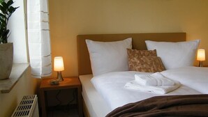 Habitación de hotel con una cama, dos lámparas de noche, una mesita de noche, una planta en maceta y un ventanal con cortinas.