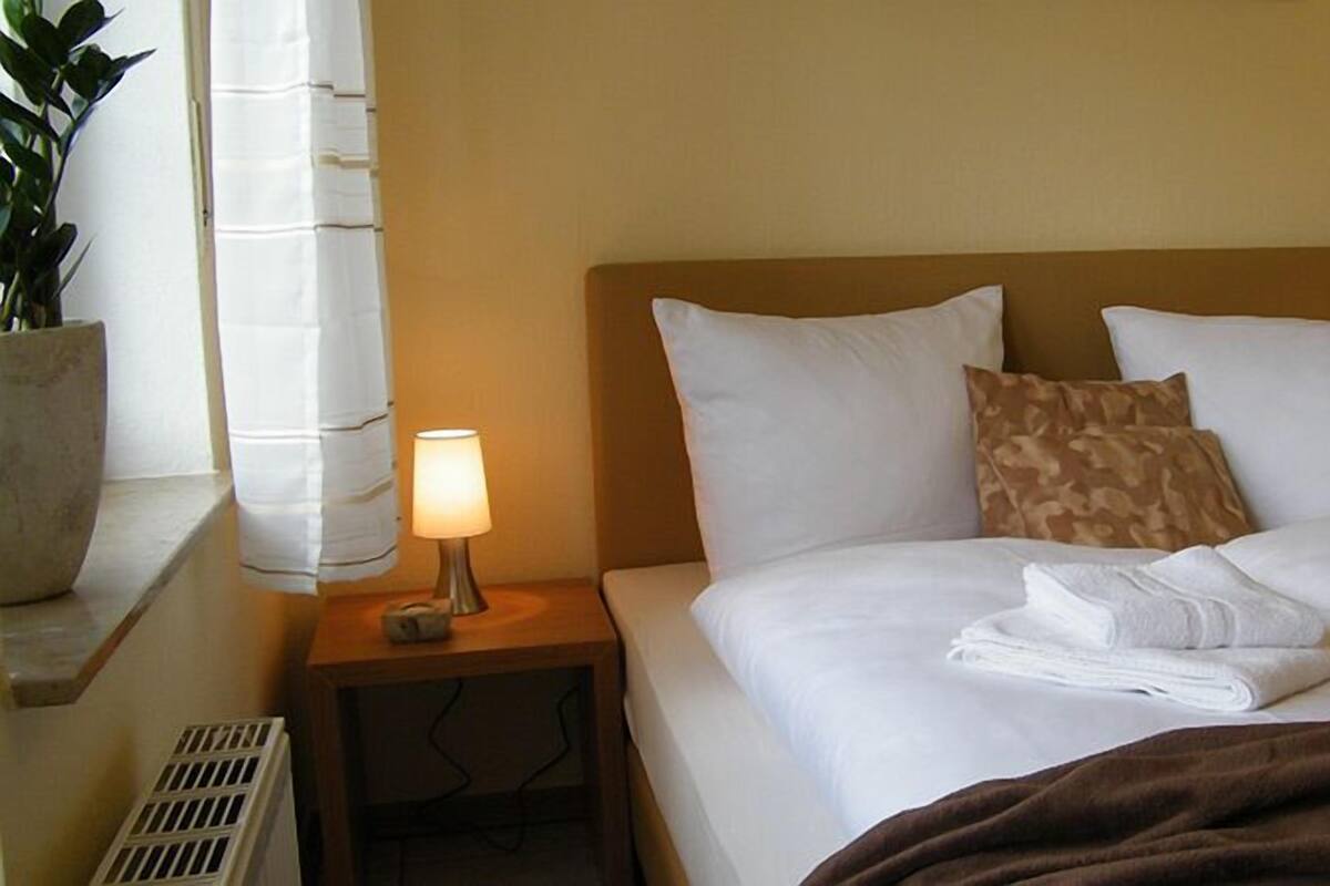 Ein Hotelzimmer mit einem Bett, zwei Nachttischlampen, einem Nachttisch, einer Topfpflanze und einem Fenster mit Vorhängen.