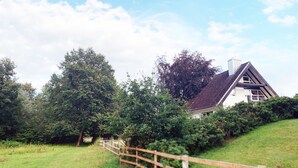 A house with a steeply pitched roof, surrounded by lush greenery and a wooden fence.