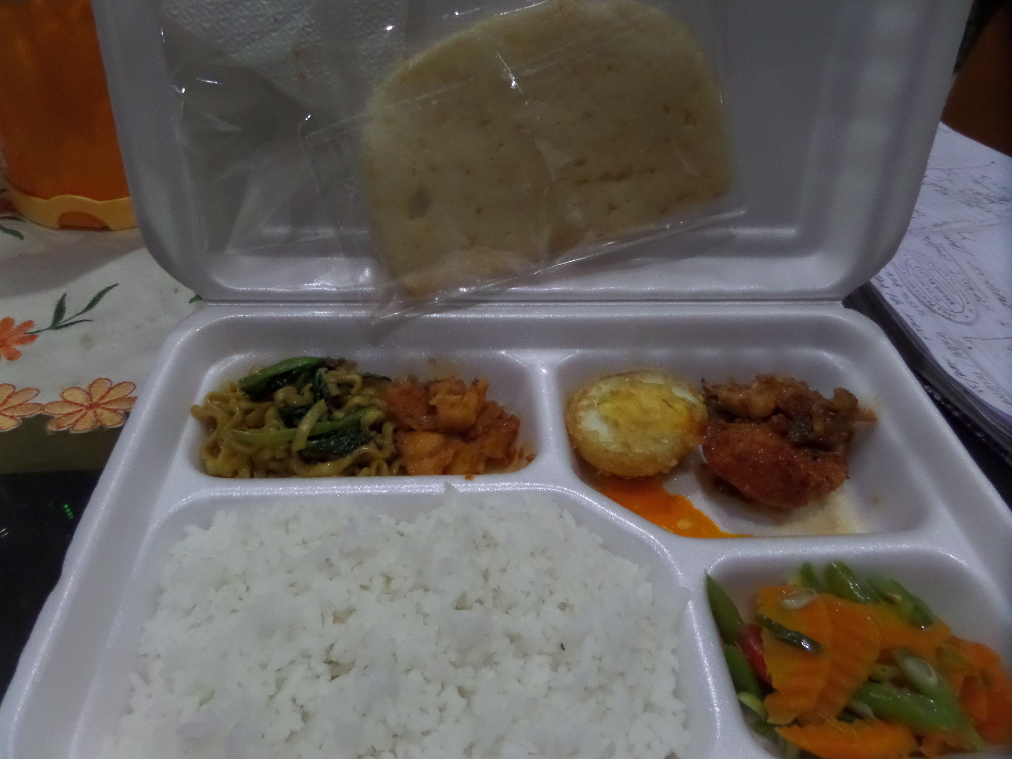 A takeout meal with rice, vegetables, and a bread roll in a styrofoam container.