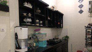 Uma cozinha compacta com armários pretos, uma geladeira branca e um fogão a gás. Há prateleiras sobre o balcão com diversos utensílios de cozinha e uma pequena planta.