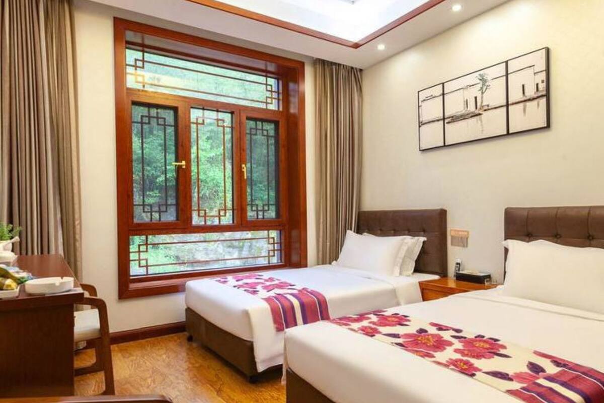 Ein Hotelzimmer mit zwei Betten, einem Holztisch, einem großen Fenster mit Vorhängen und einem an der Wand befestigten Kunstwerk.