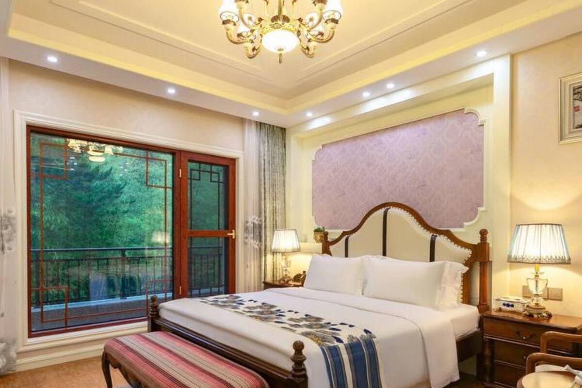 Ein Schlafzimmer mit einem großen Bett, einer Bank, einem Nachttisch, einem Sessel und einem Fenster mit Blick auf Grünflächen.