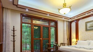 A hotel room with a wooden floor, a bed with a white bedspread, a bench, a desk, and a chair. There is a large window with a view of greenery outside. - Floral Hotel Enshi Xiaoxihu Yihao (Enshi)