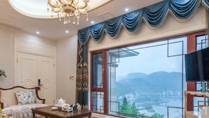 A spacious room with a large window offering a view of a river and mountains. There is a chandelier, a wooden table, and a sofa with a patterned cover. - Floral Hotel Enshi Xiaoxihu Yihao (Enshi)