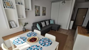 Een moderne woonkamer met een grijze hoekbank, een eettafel met geruite placemats, en een keuken met een koelkast en kasten.