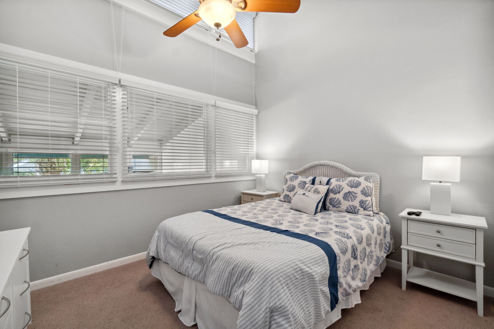 A bedroom with a bed, nightstands, a ceiling fan, and large windows with blinds.