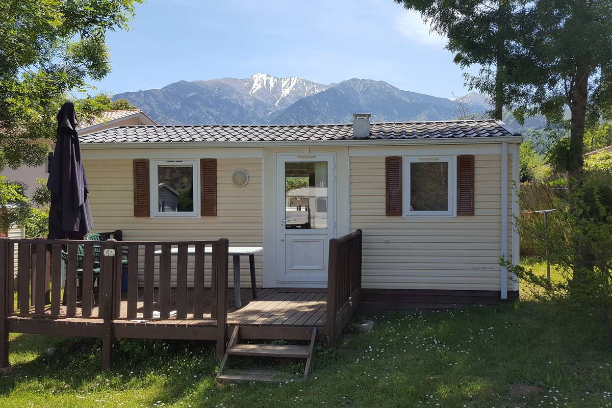 A small, single-story mobile home with a front porch, a dining table, and a chair. It features a window with shutters and a door.