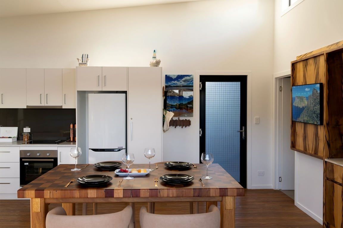 A modern kitchen with white cabinets, a wooden dining table set for four, and a refrigerator.