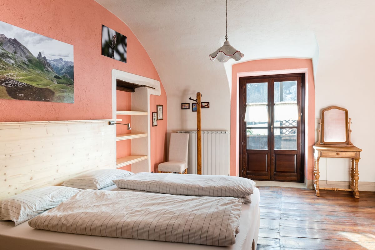 A bedroom with a bed, a wooden headboard, a radiator, a chair, a wooden table, and a door with a view outside.