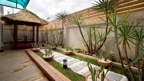 Una terraza de madera con techo de paja, rodeada de plantas en macetas y un sendero de piedra.