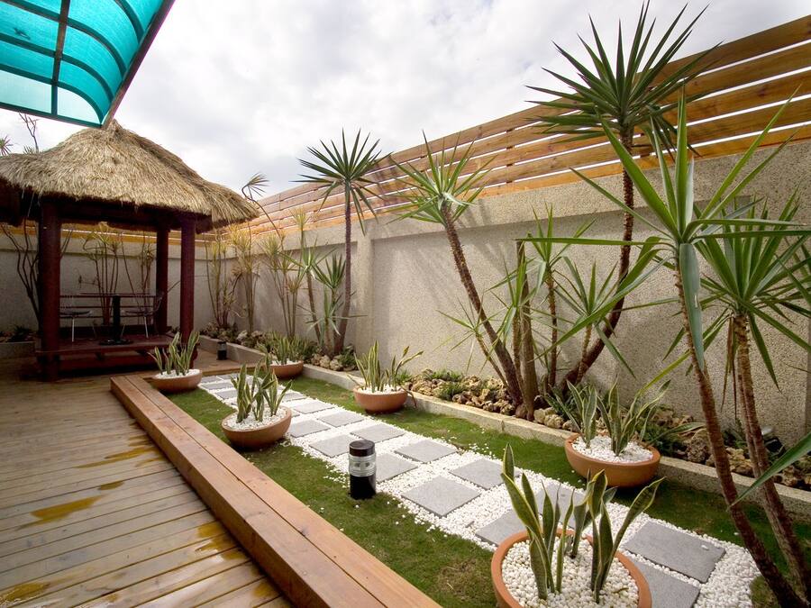 Una terraza de madera con techo de paja, rodeada de plantas en macetas y un sendero de piedra.