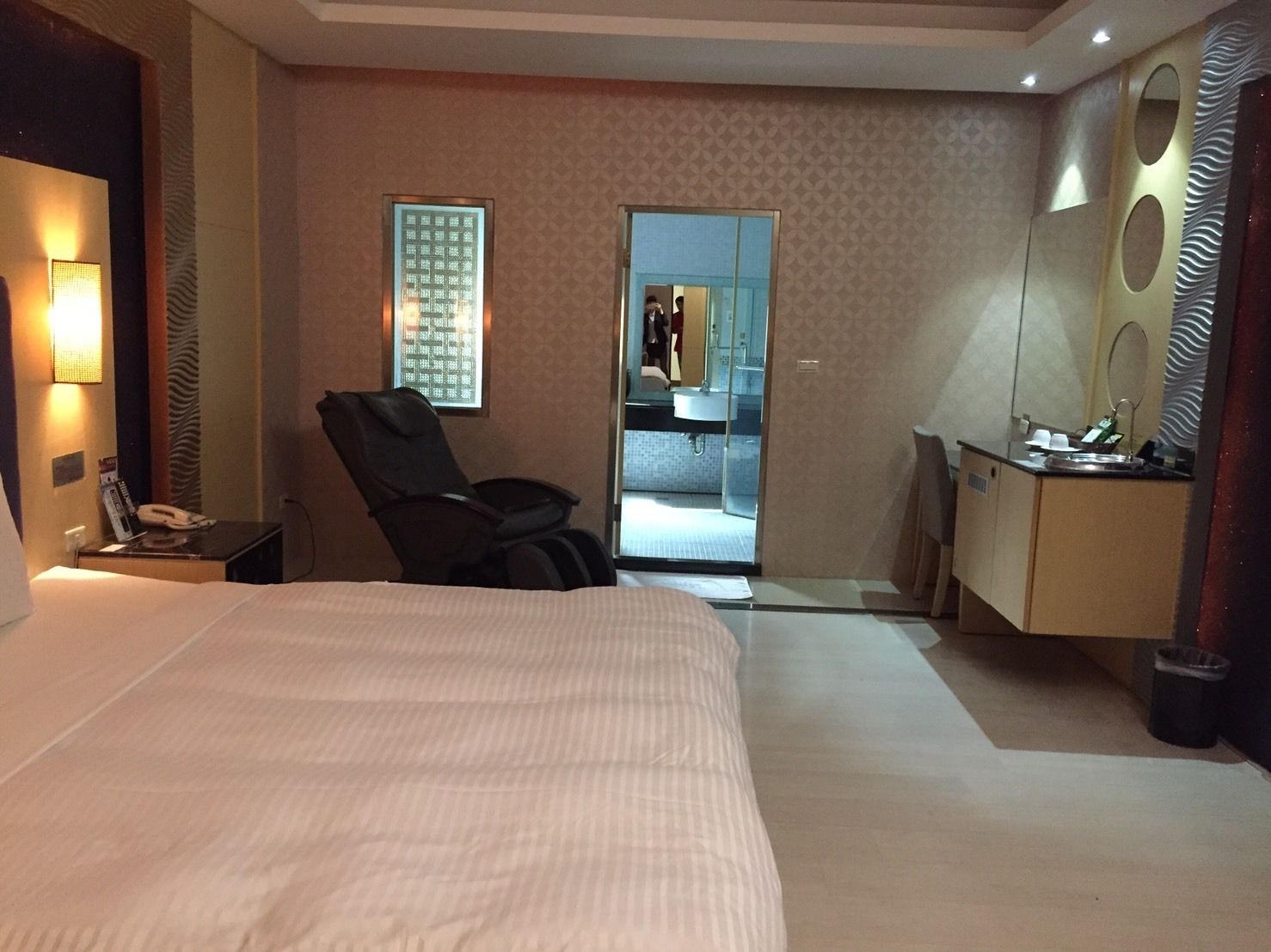 A hotel room with a large bed, a massage chair, a desk with a mirror, and a bathroom visible through an open door.