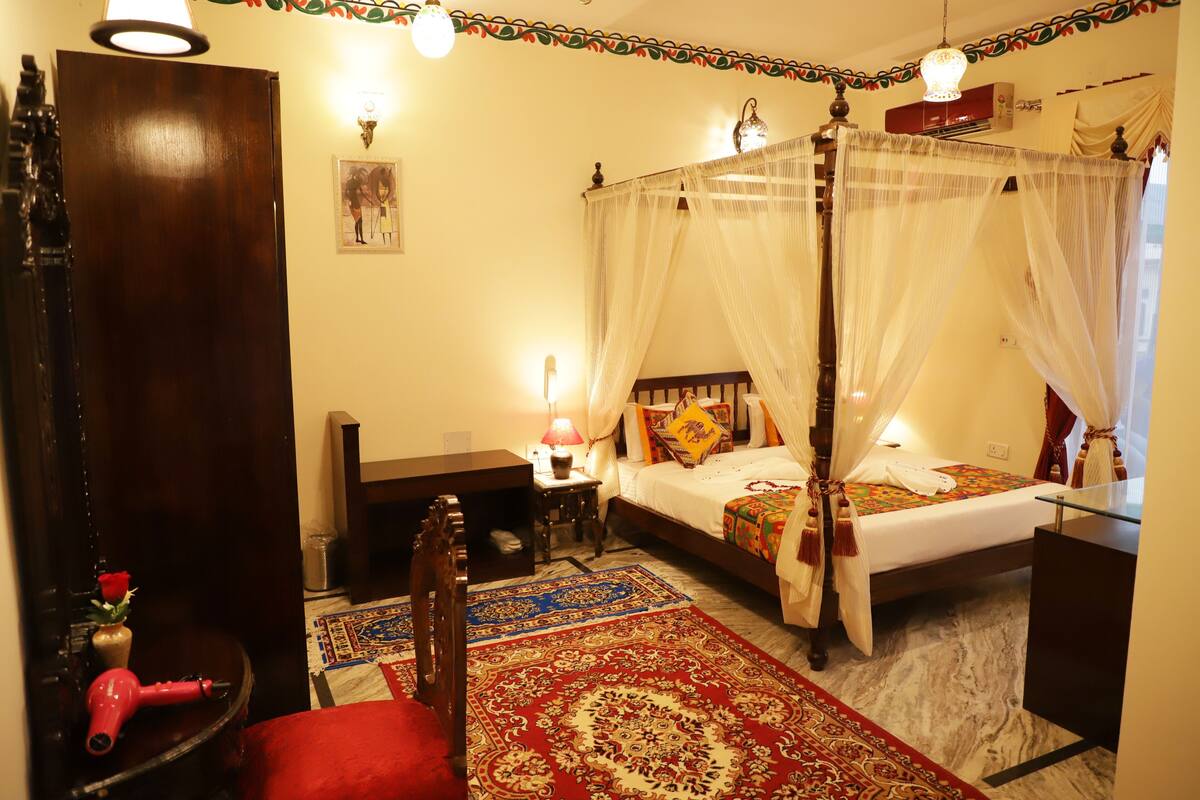 A traditional four-poster bed with a canopy, a red chair, a wooden desk, and a small table with a lamp.