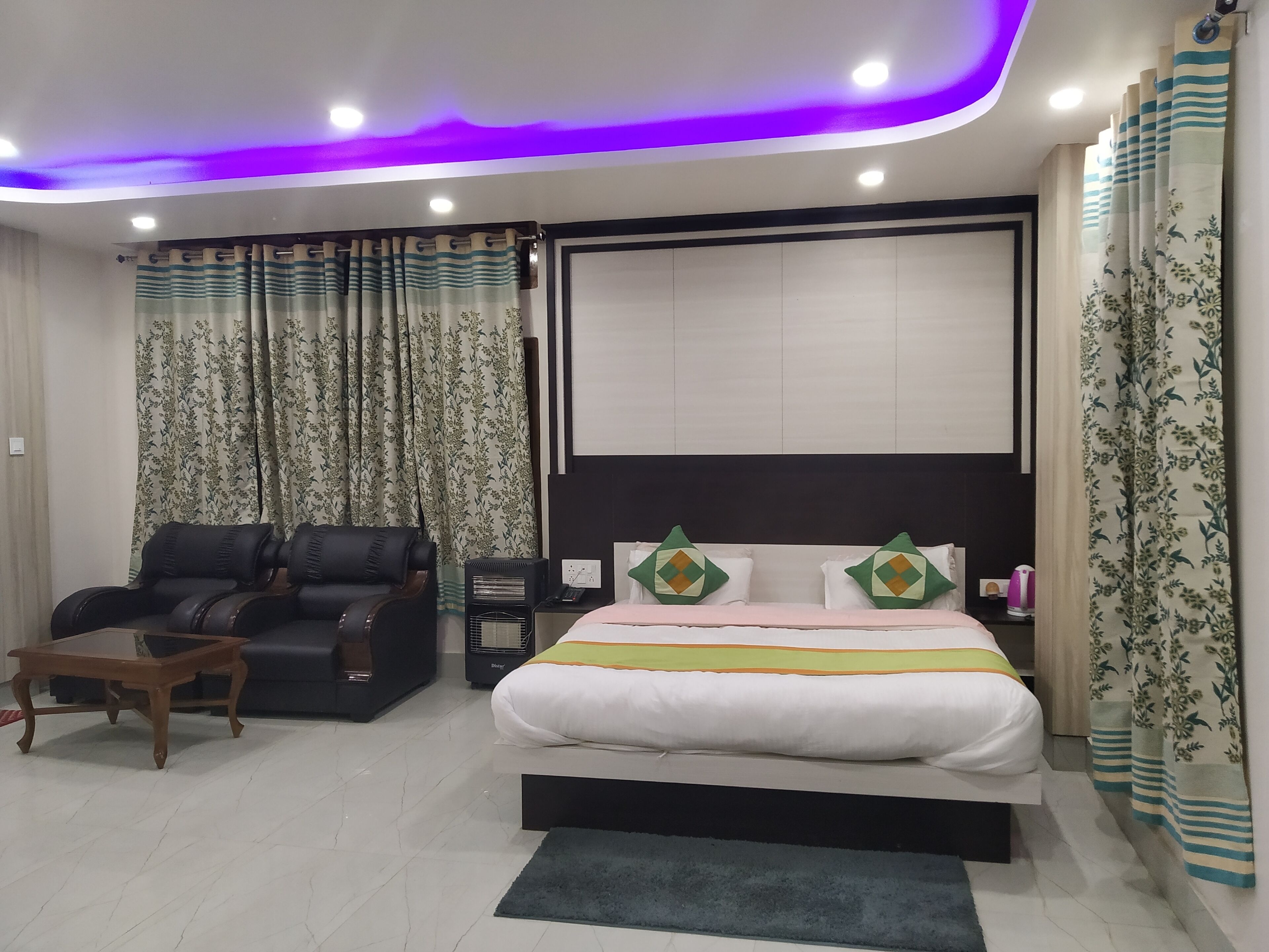 Una habitación de hotel moderna con una cama grande, un sofá, una mesita pequeña e iluminación morada en el techo.