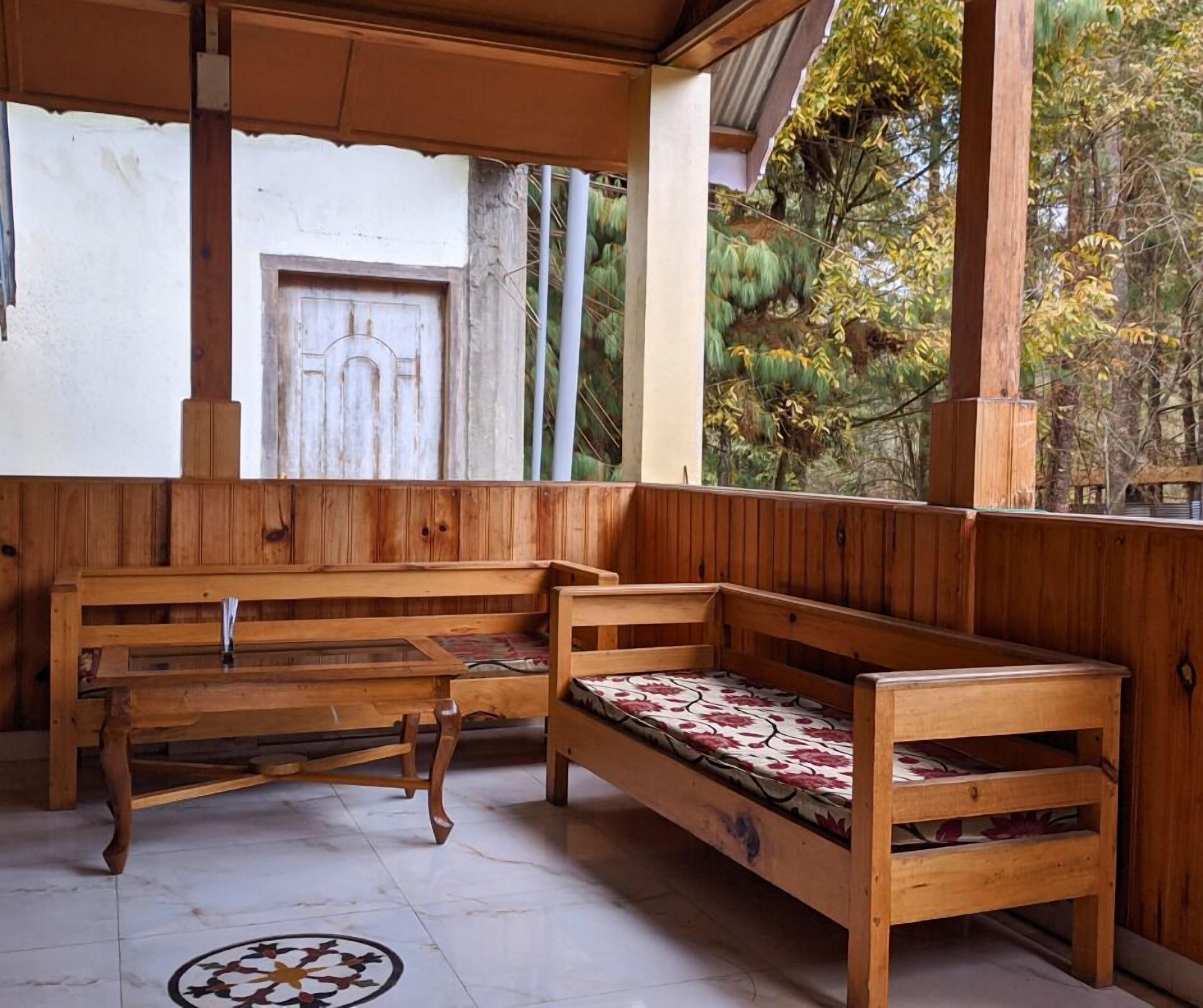 A wooden bench with a patterned cushion in a covered outdoor area with a view of trees and a building in the background.