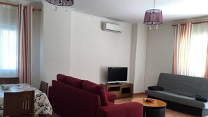 Un salon avec un canapé rouge, un canapé gris, un téléviseur posé sur un meuble en bois, une table à manger avec des chaises et un lustre.