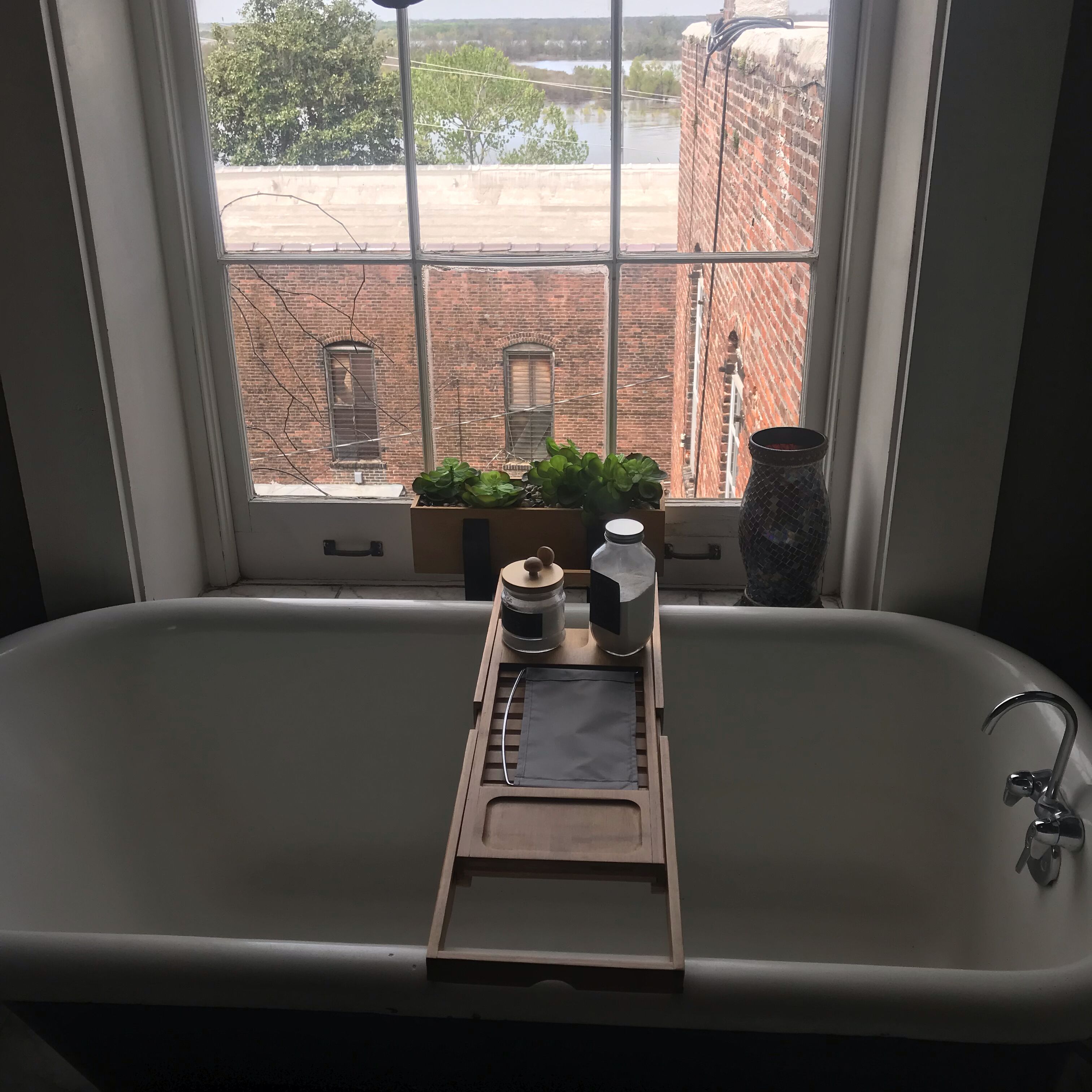 A bathroom with a window, a sink, a wooden tray, a potted plant, and a vase.