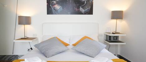 Habitación de hotel con cama, dos mesitas de noche, vista panorámica de la ciudad y un reloj digital en la mesita.