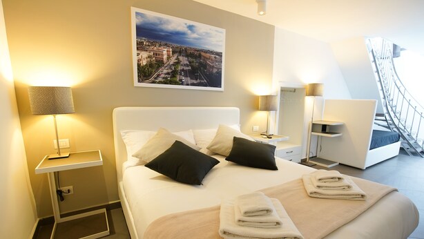 A hotel room with a neatly made bed, two bedside lamps, a cityscape painting, and a view of the outside balcony. - Le Stanze di Piazza Cairoli (Messina)
