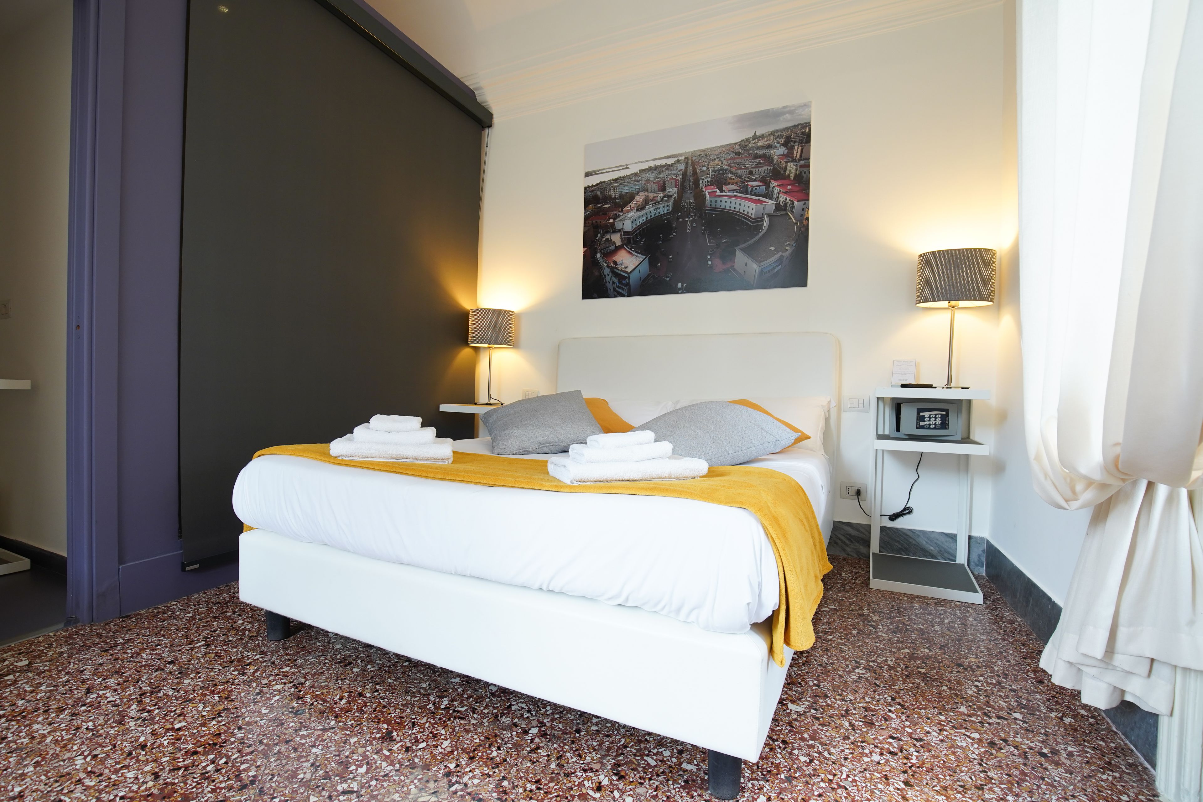 Une chambre d’hôtel avec un lit, une table de chevet avec une lampe, un tableau au mur et une vue sur le paysage urbain.