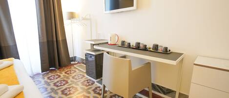 A hotel room with a bed, a desk with a kettle, a TV, and a small refrigerator.
