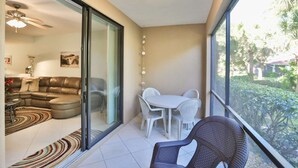 A living room with a sofa, a coffee table, and a dining area with a table and chairs. There is a sliding glass door leading to a balcony with a view of greenery.
