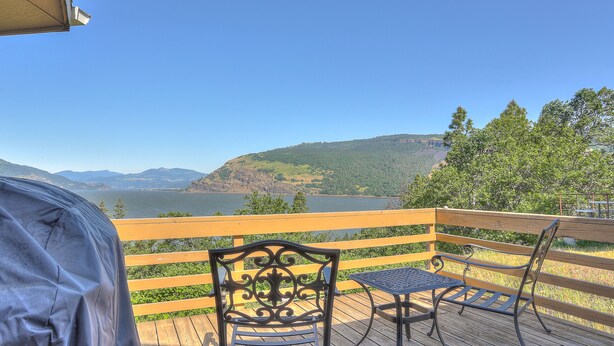 Eine Holzterrasse mit einem Metalltisch und Stühlen, mit Blick auf einen See und Berge.